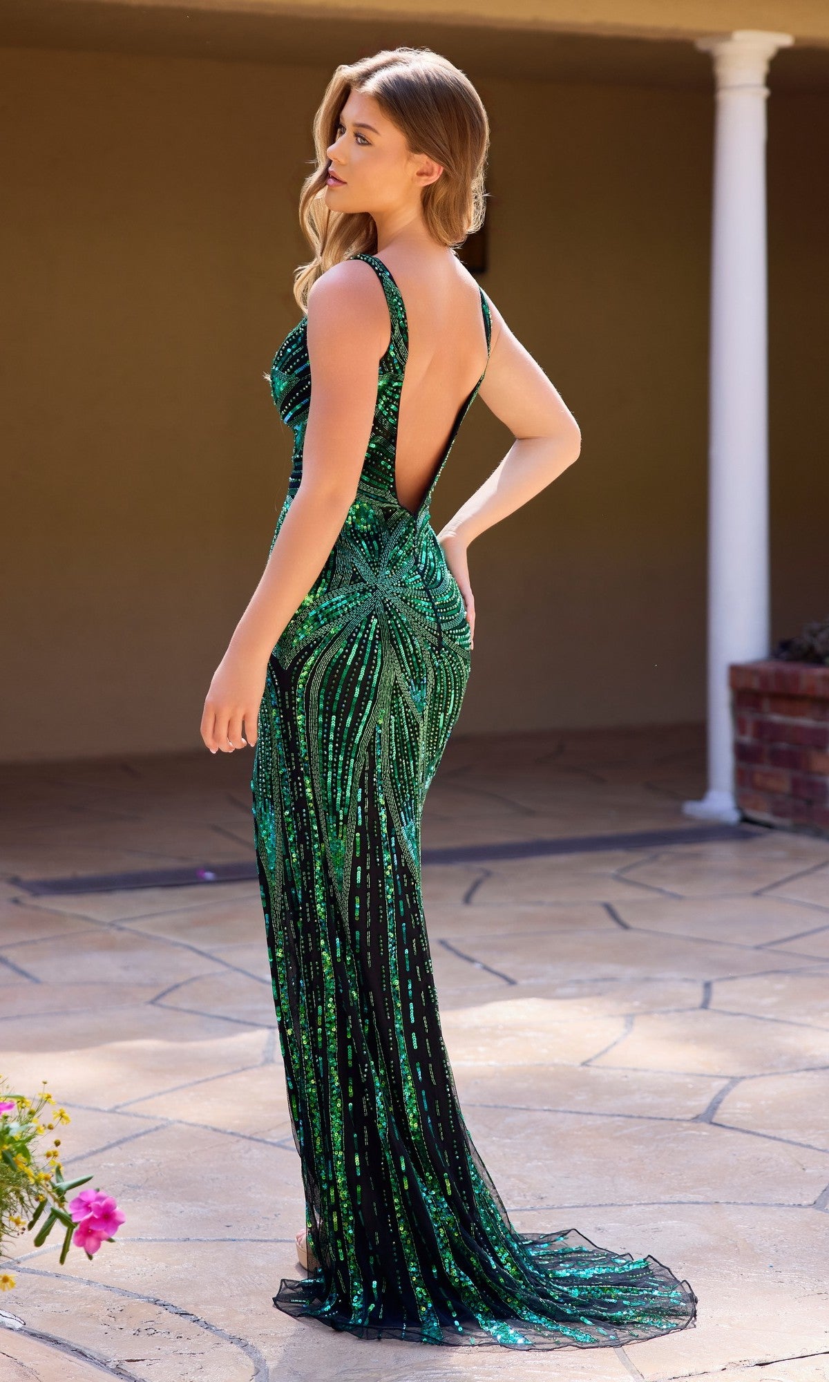 A woman with long hair stands outdoors wearing the Primavera 4502, a fitted sleeveless green sequin prom gown with a low back and slight train.